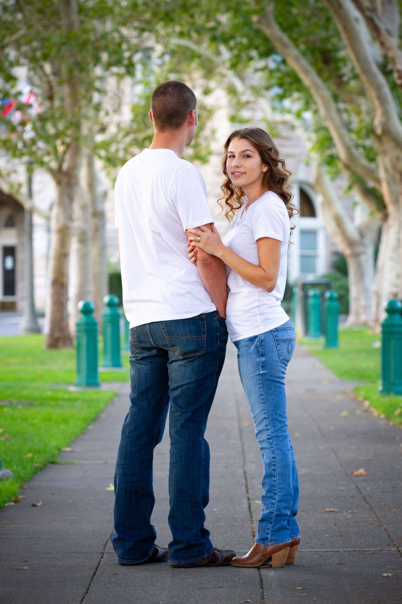 Casual couple in white shirts and jeans dancing on tree-lined pathway.