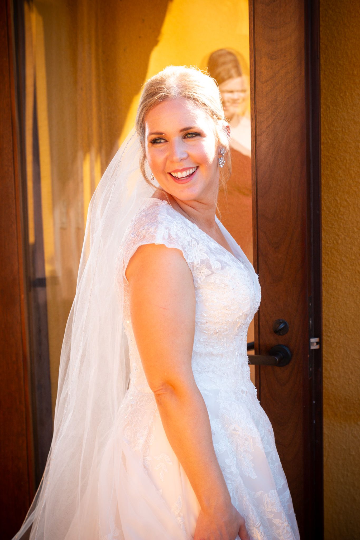 Bride in white lace wedding dress smiles brightly against warm orange wall background.