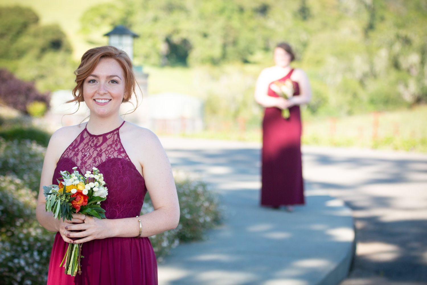Bridesmaid in burgundy dress holds colorful bouquet outdoors with another attendant visible in background.