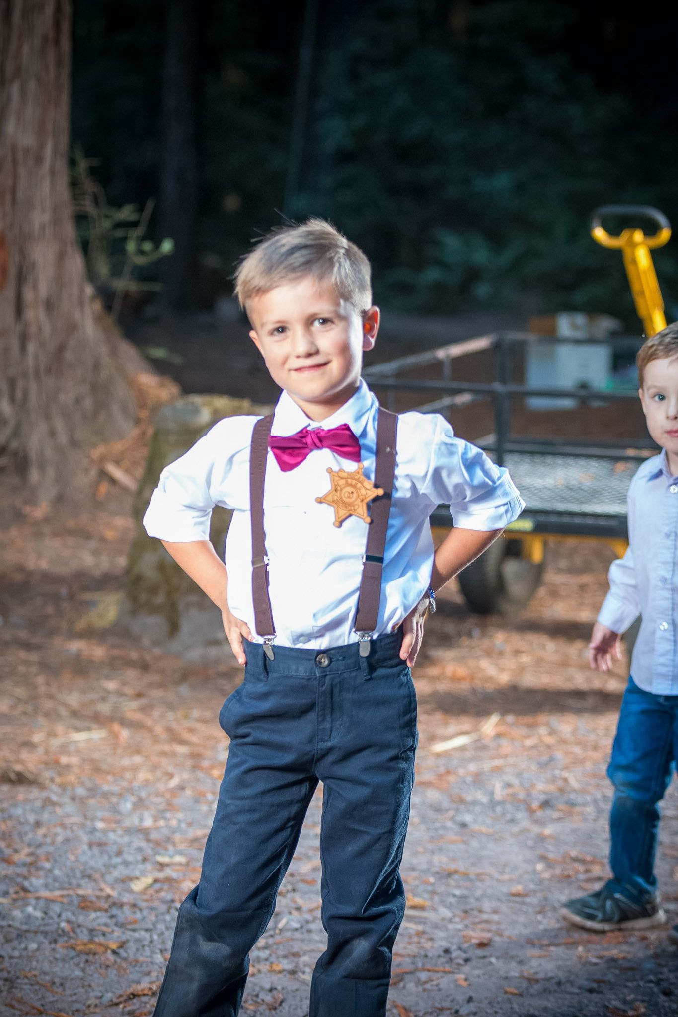 Ring bearer in suspenders and bow tie poses confidently at outdoor wedding.