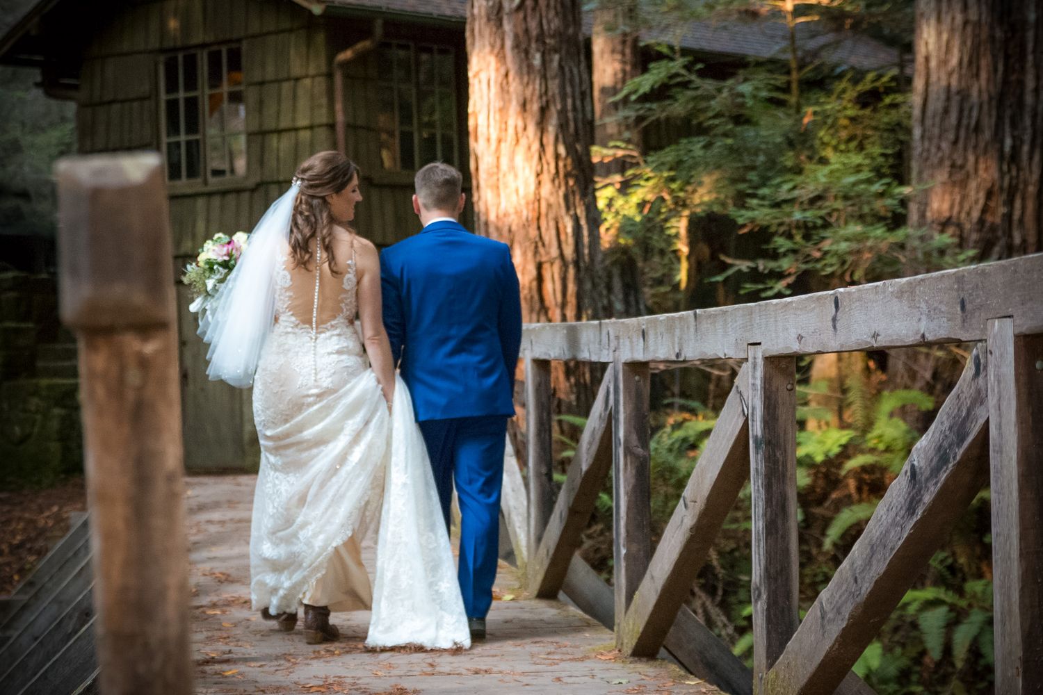 Newlyweds walk hand in hand across a wooden bridge with a rustic cabin visible in the background.