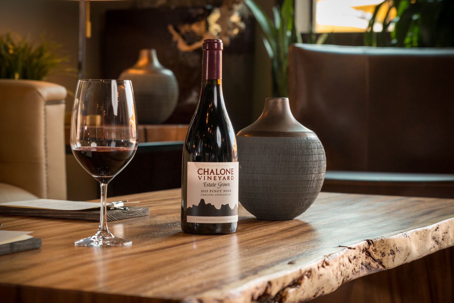 Wine bottle and glass staged on live edge table with decorative ceramic vase and ambient lighting.