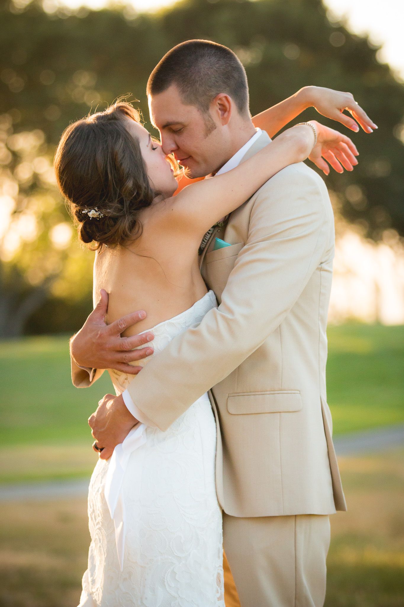 A romantic wedding couple embraces in warm golden sunlight outdoors while wearing cream colored attire.