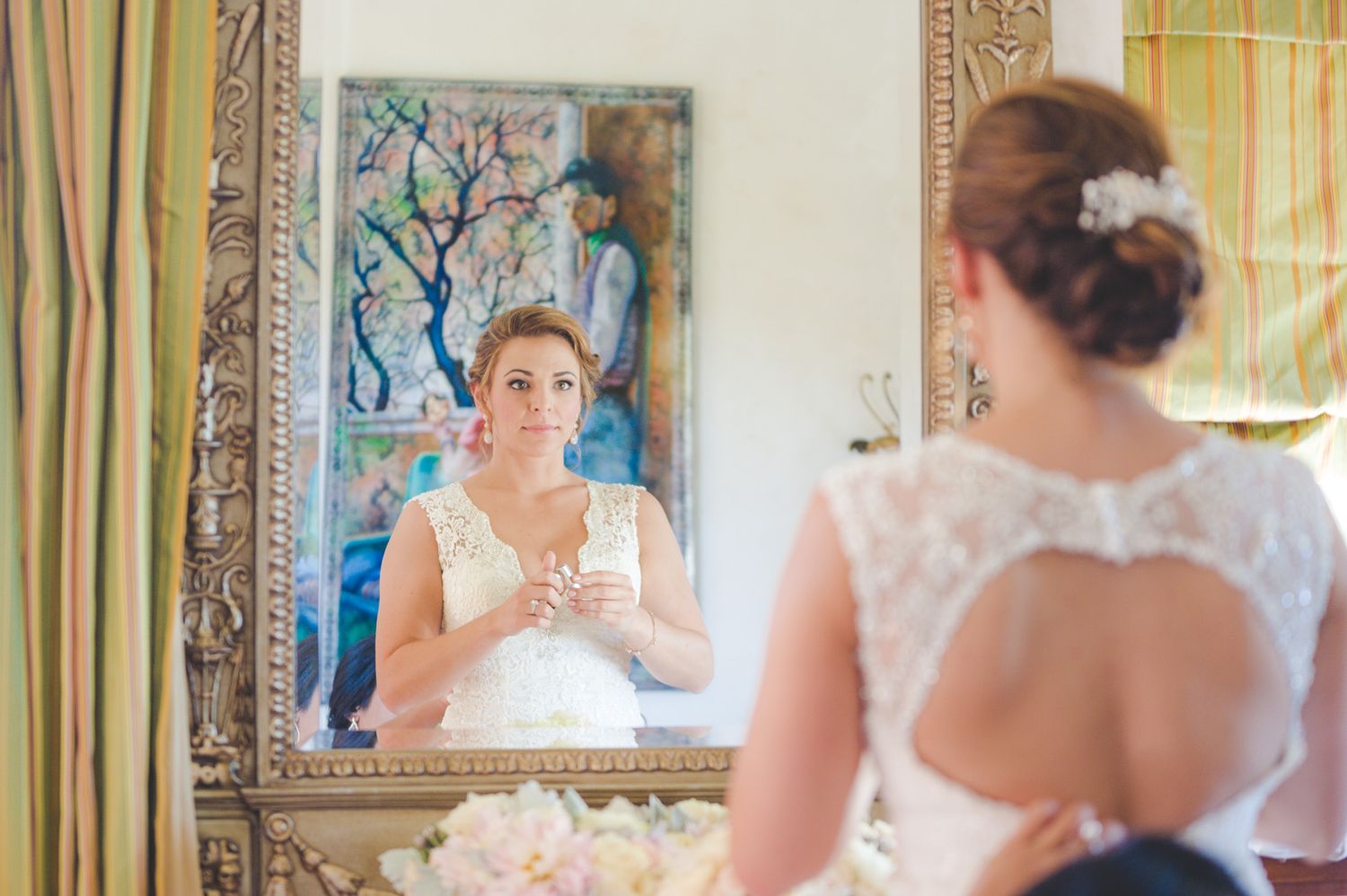 Bride in elegant lace keyhole back wedding dress holding bouquet while looking in ornate mirror reflection.