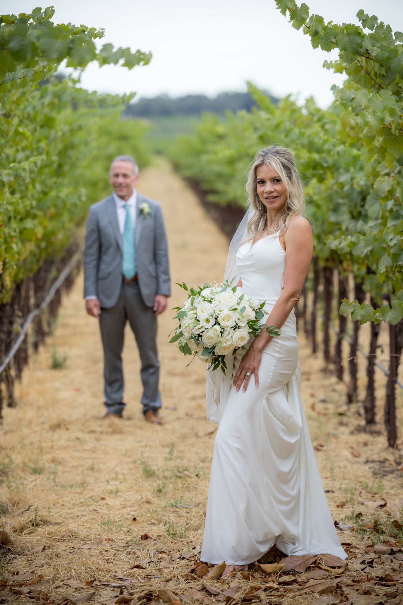 Bride with bouquet poses in vineyard with groom in background.