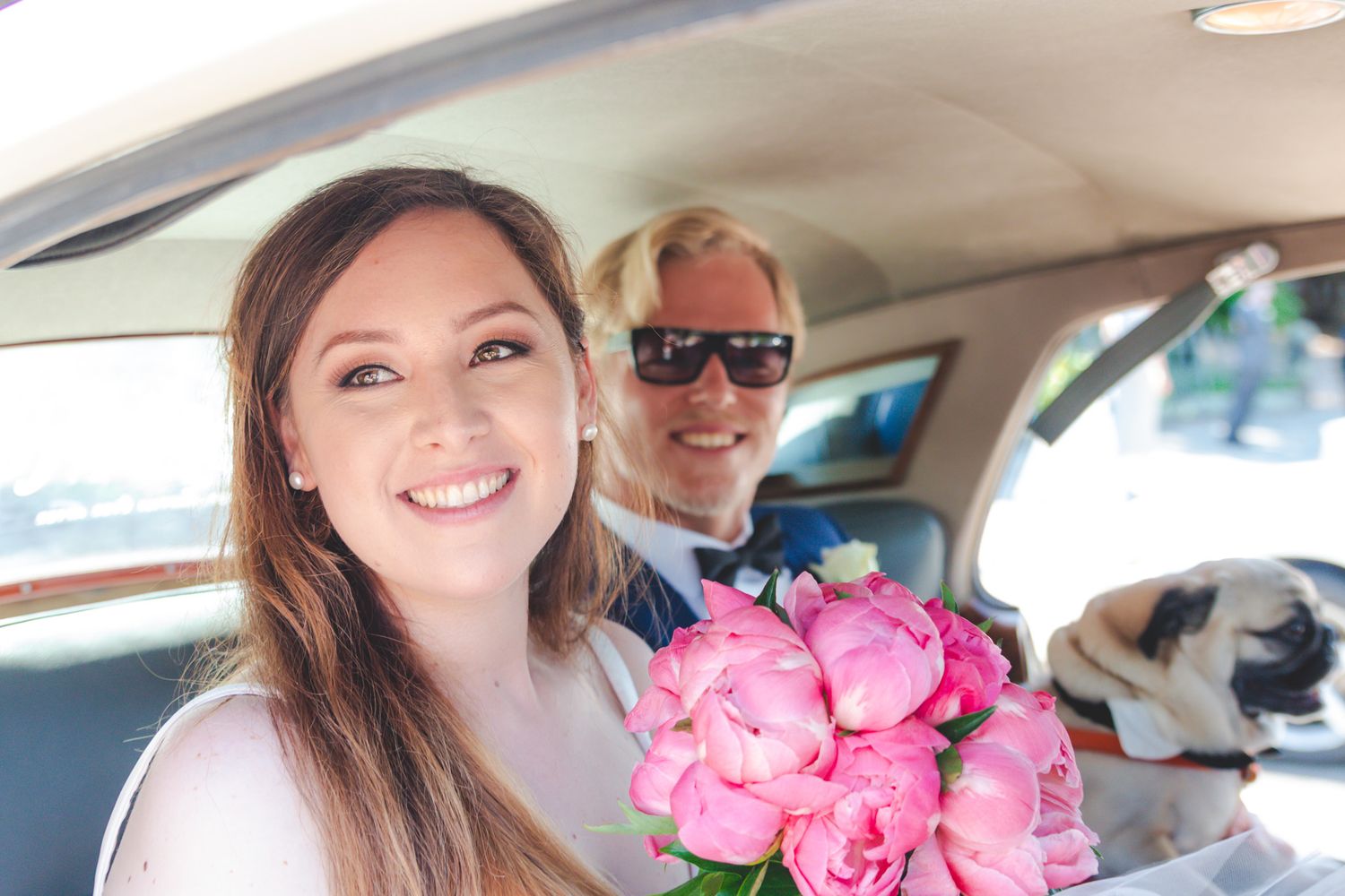 A smiling woman holds pink peonies in a car with another passenger.