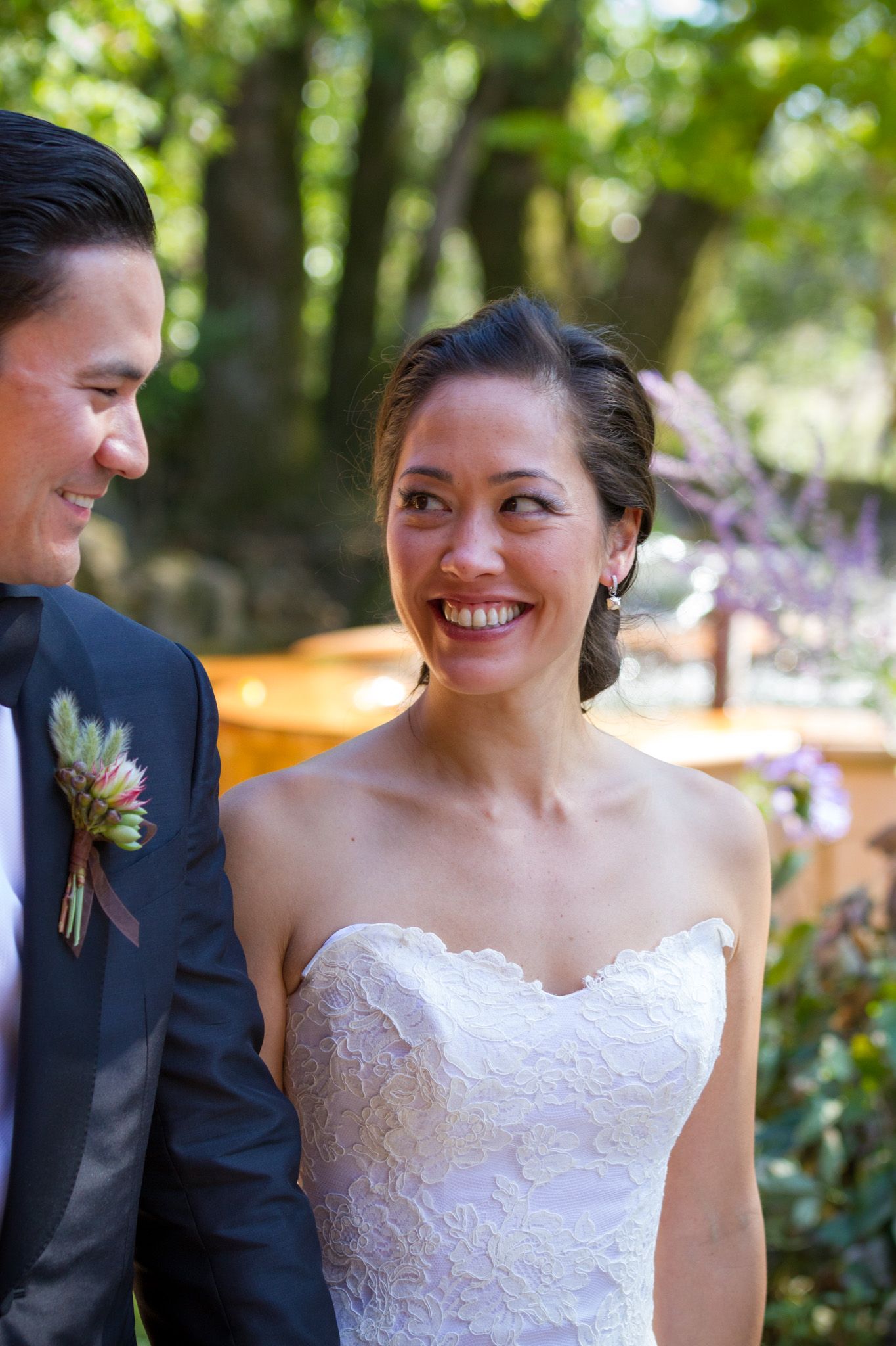 Bride in white lace dress smiling outdoors surrounded by greenery.