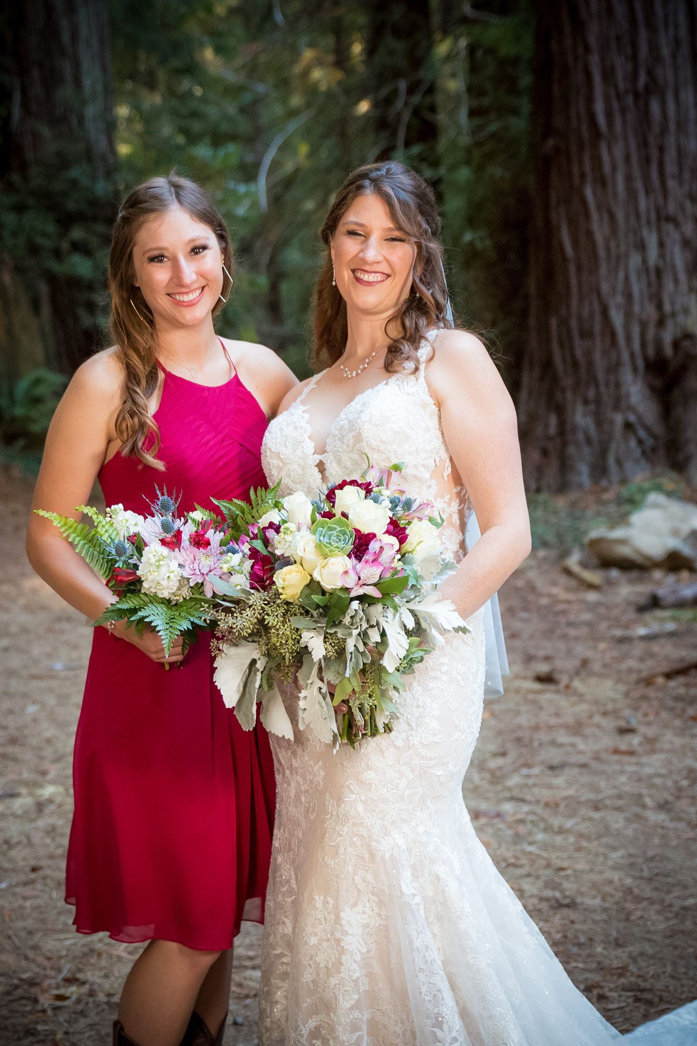 Two women holding beautiful bouquets smile together during outdoor wedding photoshoot.