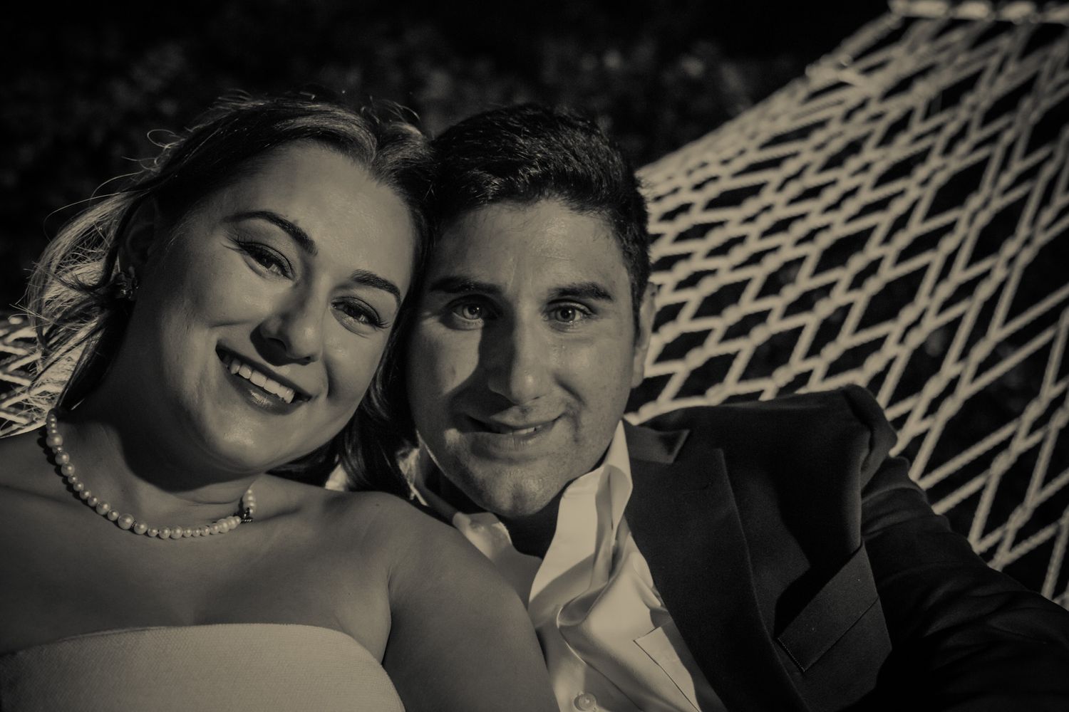 A black and white close-up portrait of a happy couple in wedding attire smiling together near a hammock.