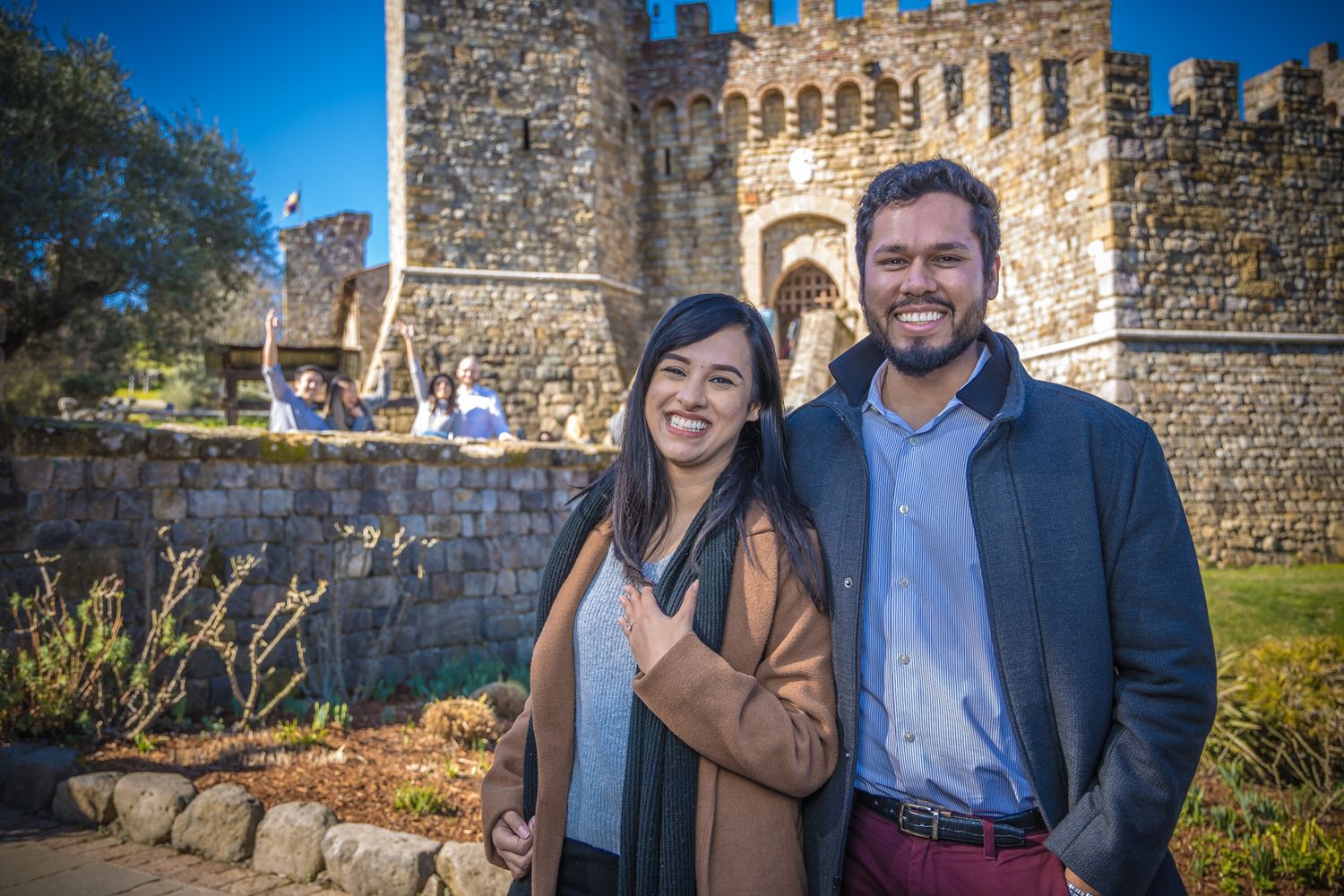 A happy couple poses together in front of a medieval castle with crenellated walls under blue sky.