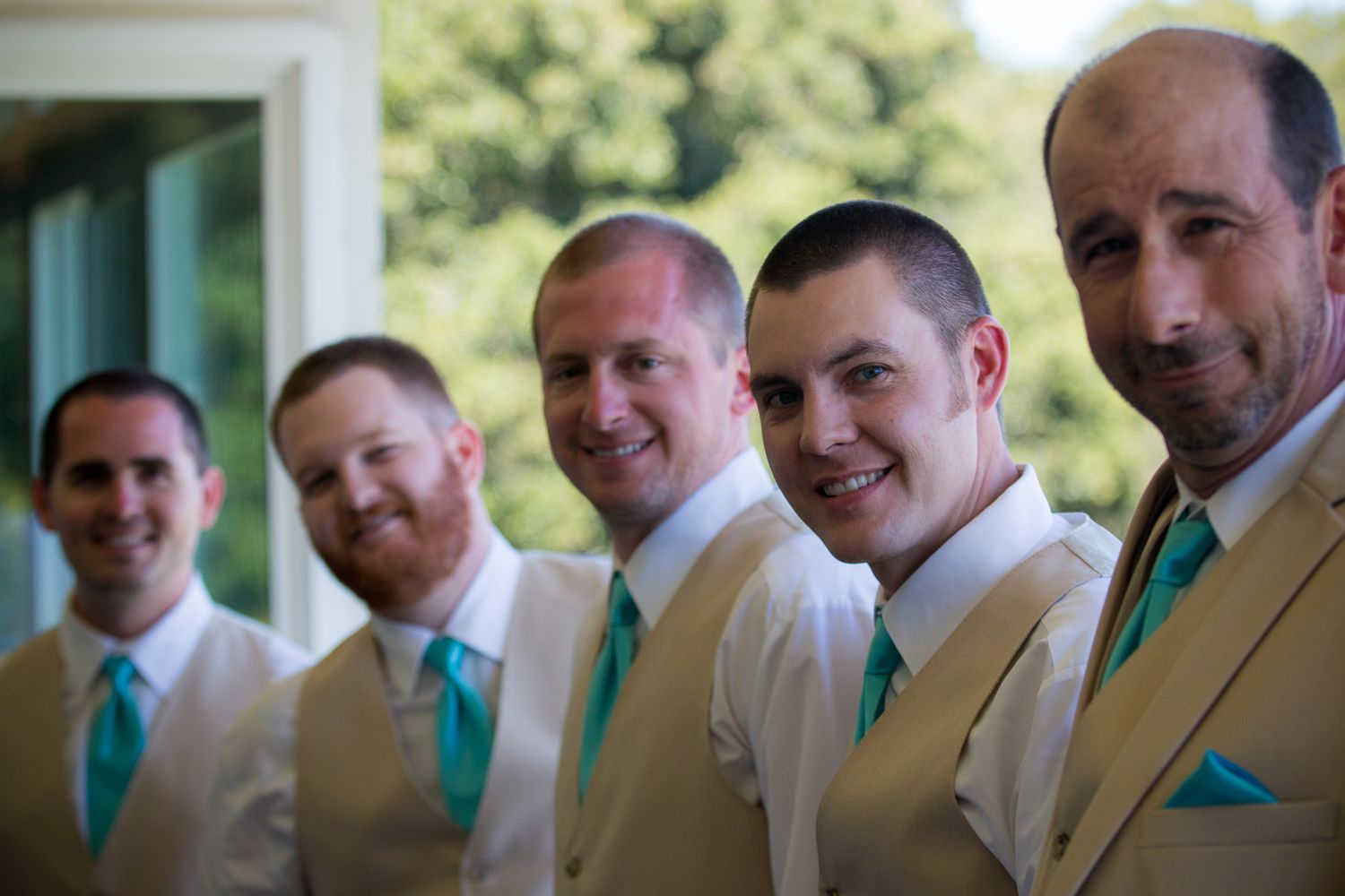 Wedding groomsmen lined up wearing white shirts and suits with matching turquoise ties and pocket squares outdoors.