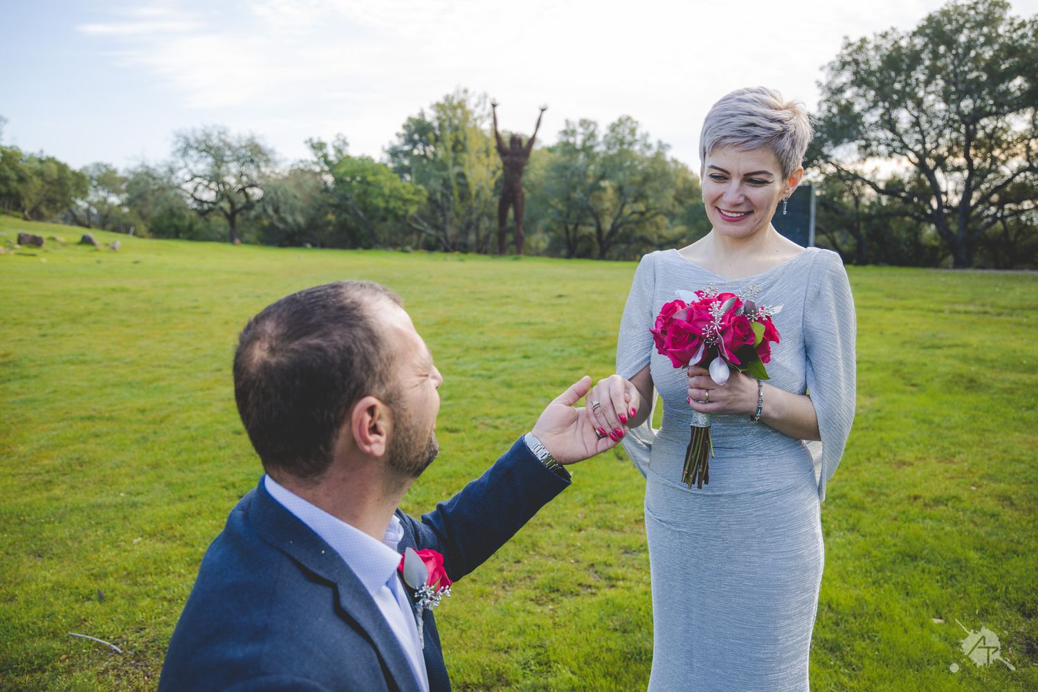 A couple in a grassy field sharing a romantic moment as one kneels and holds out their hand for the other holding flowers.
