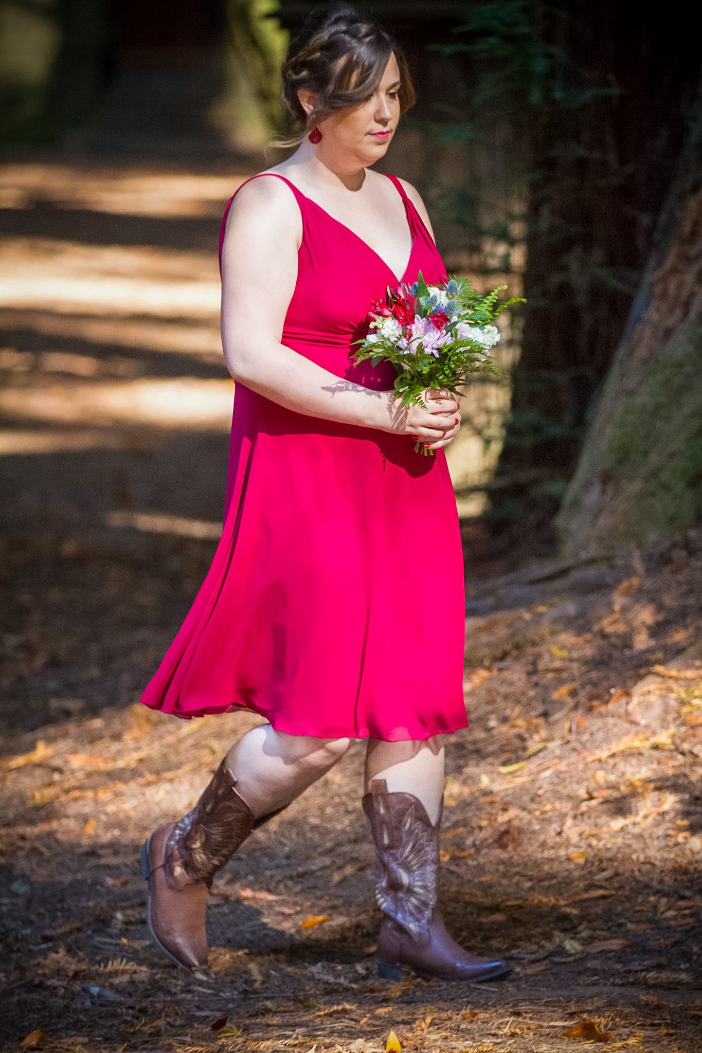 Bridesmaid in red dress holding bouquet while posing in forest setting.