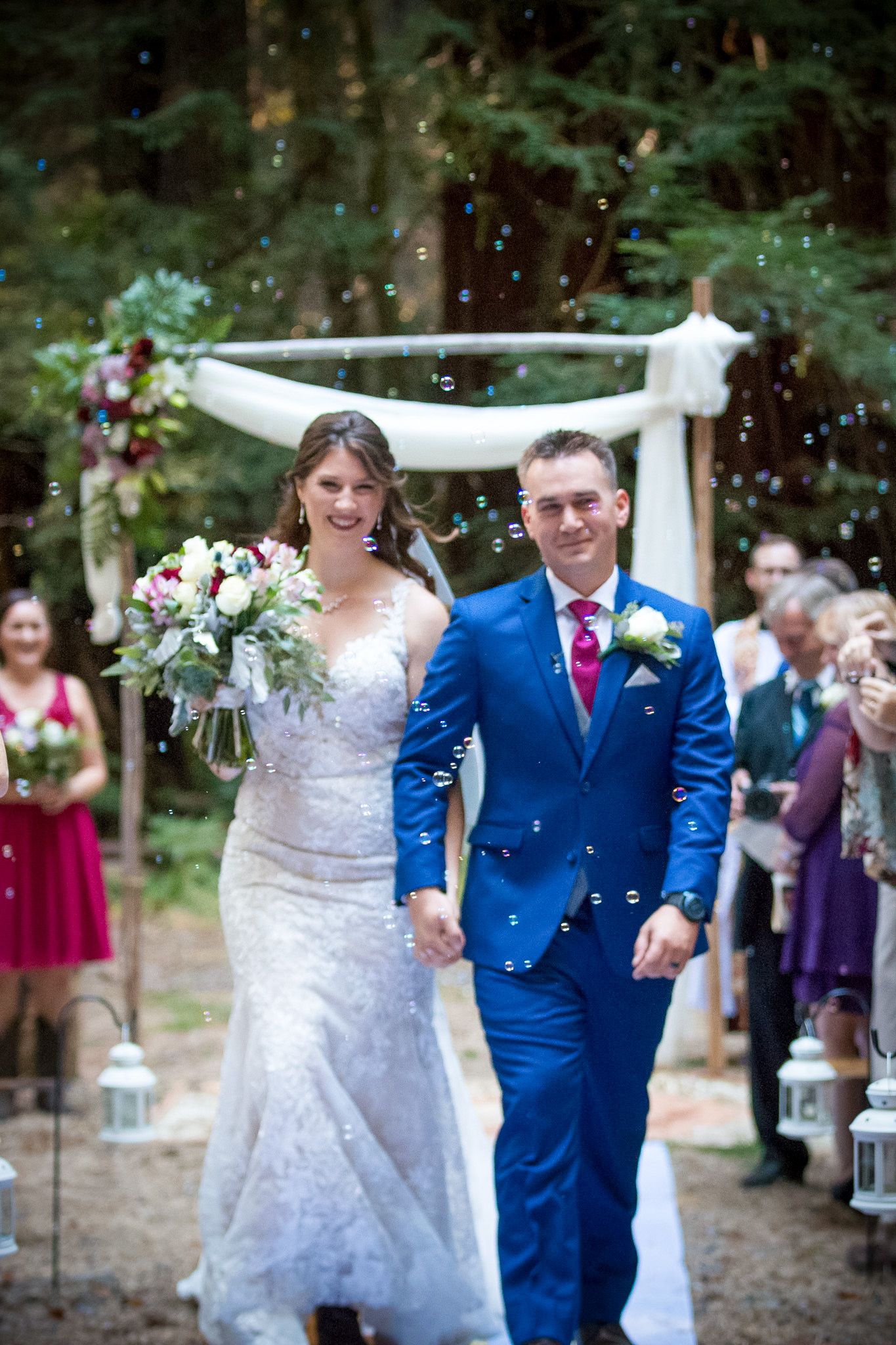 Newlyweds exit their forest ceremony under falling confetti while guests celebrate.