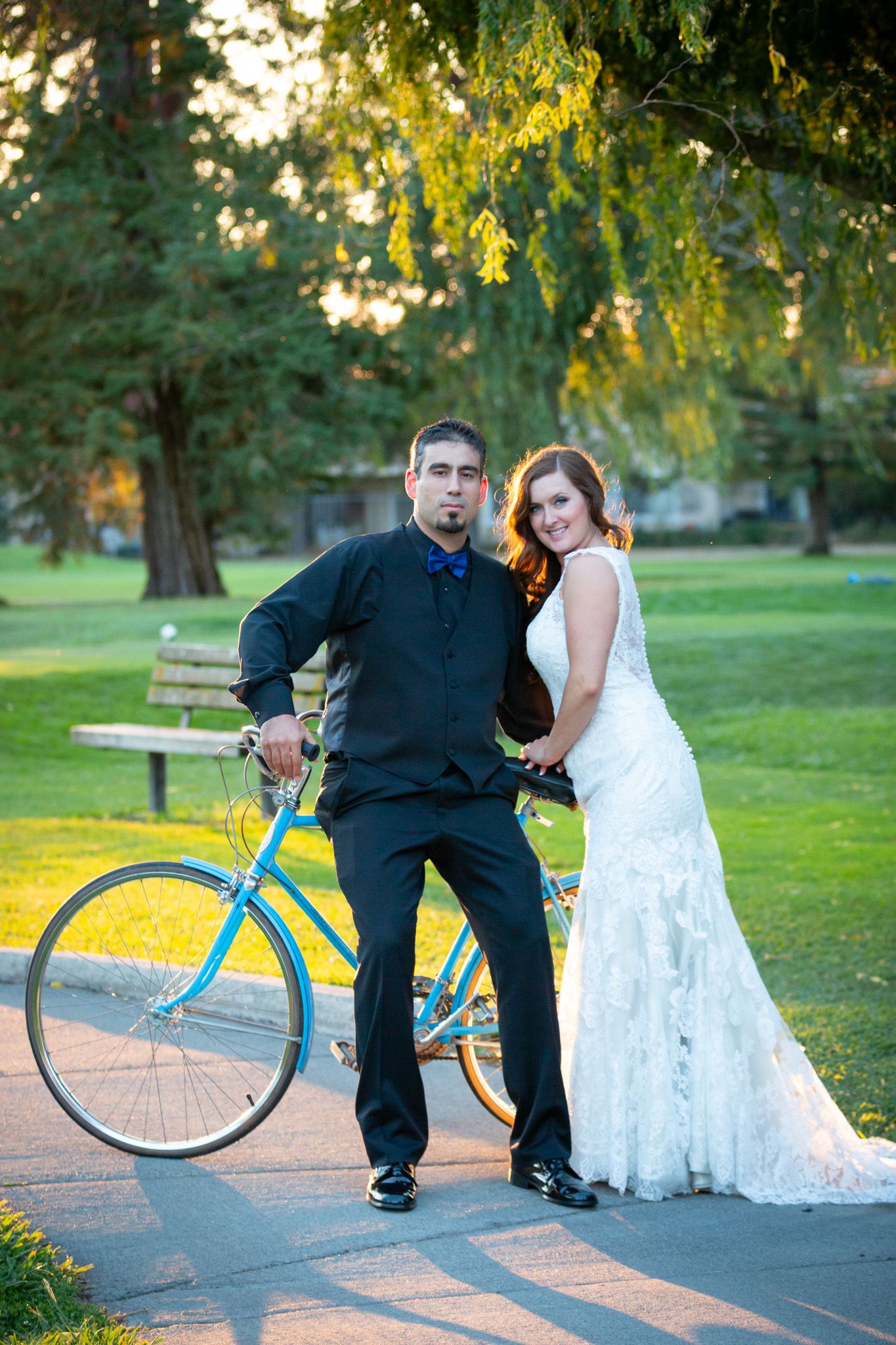 Wedding couple poses with vintage blue bicycle under hanging tree branches.