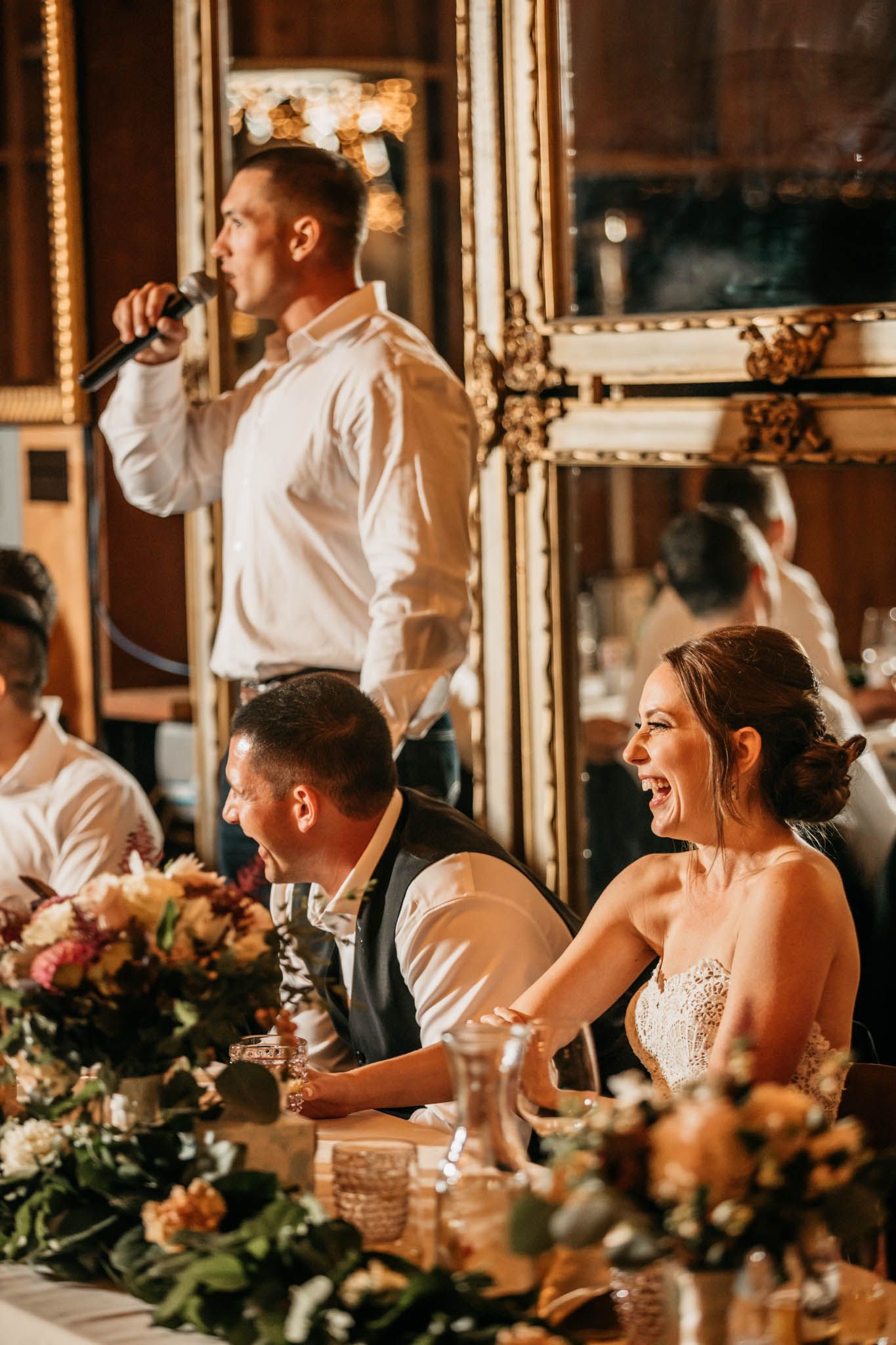 A wedding reception speech with people seated at an elegantly decorated table with floral centerpieces.