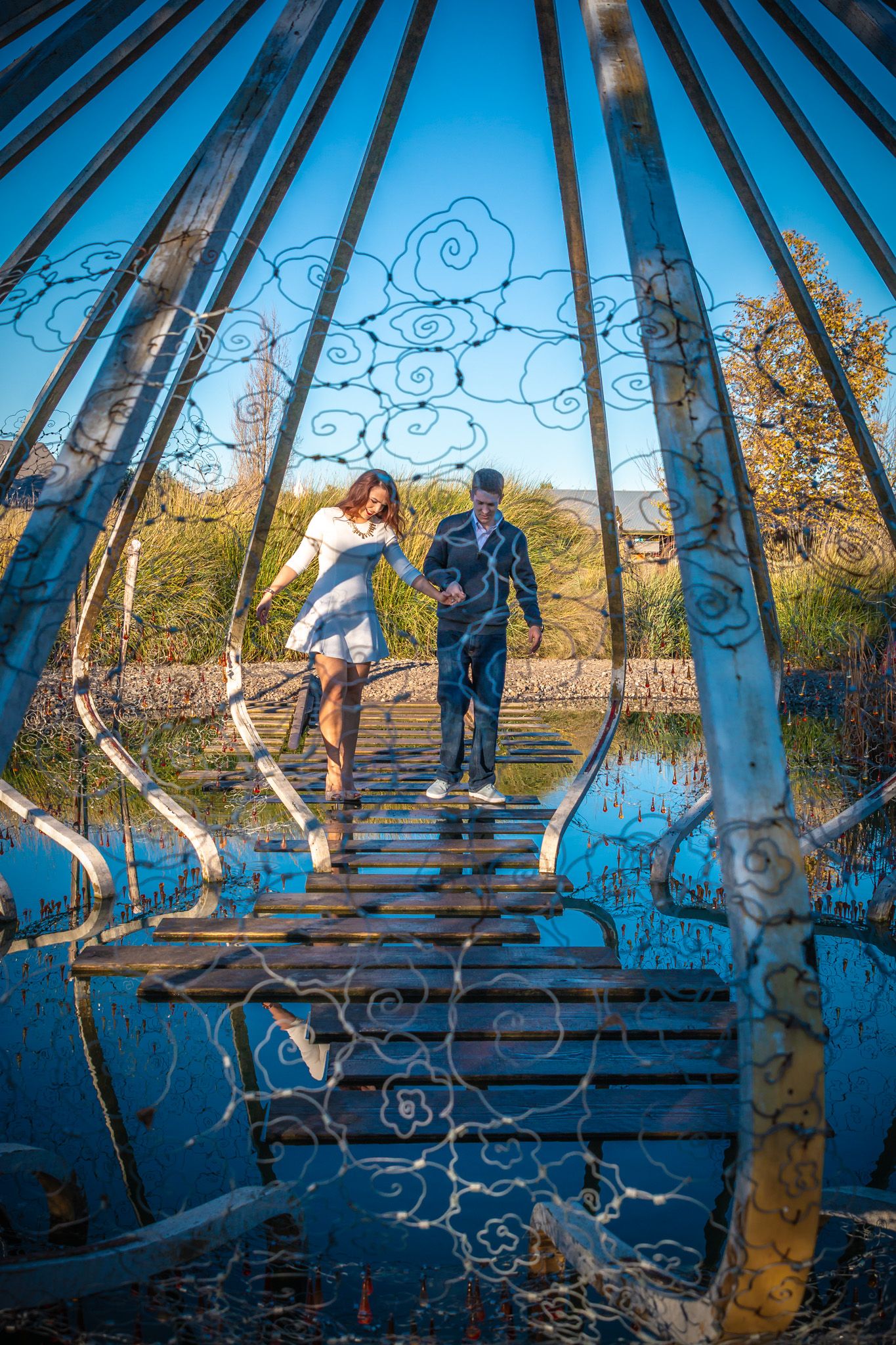 Couple walking across ornate blue bridge with decorative metalwork designs reflected in water below during daytime.