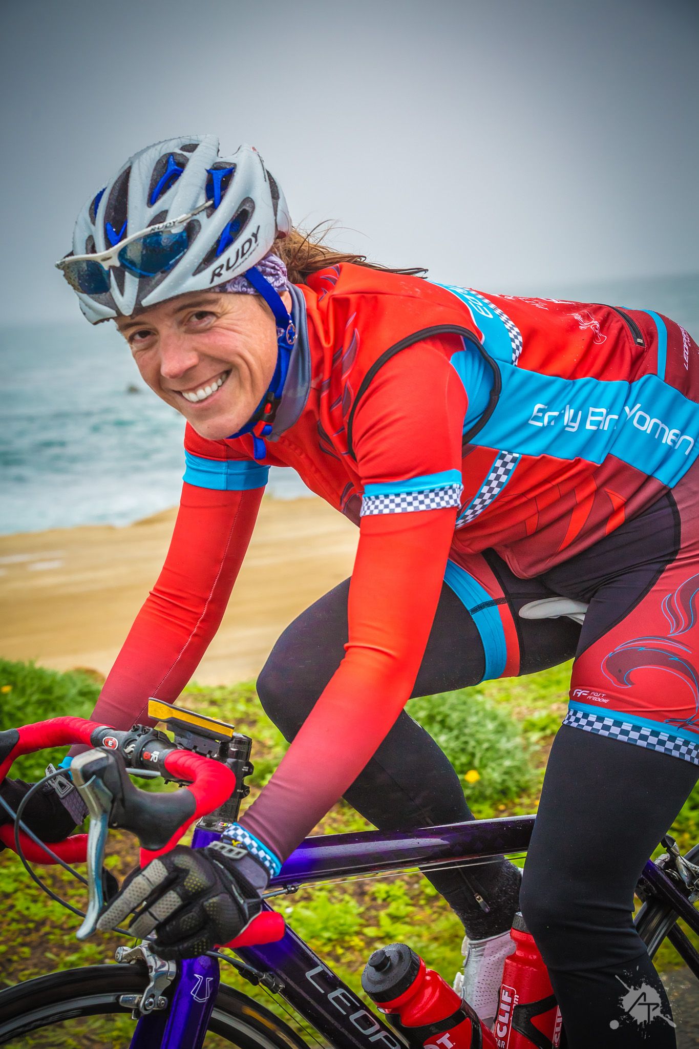 Cyclist in red and blue jersey riding a road bike along scenic coastal path on sunny day.