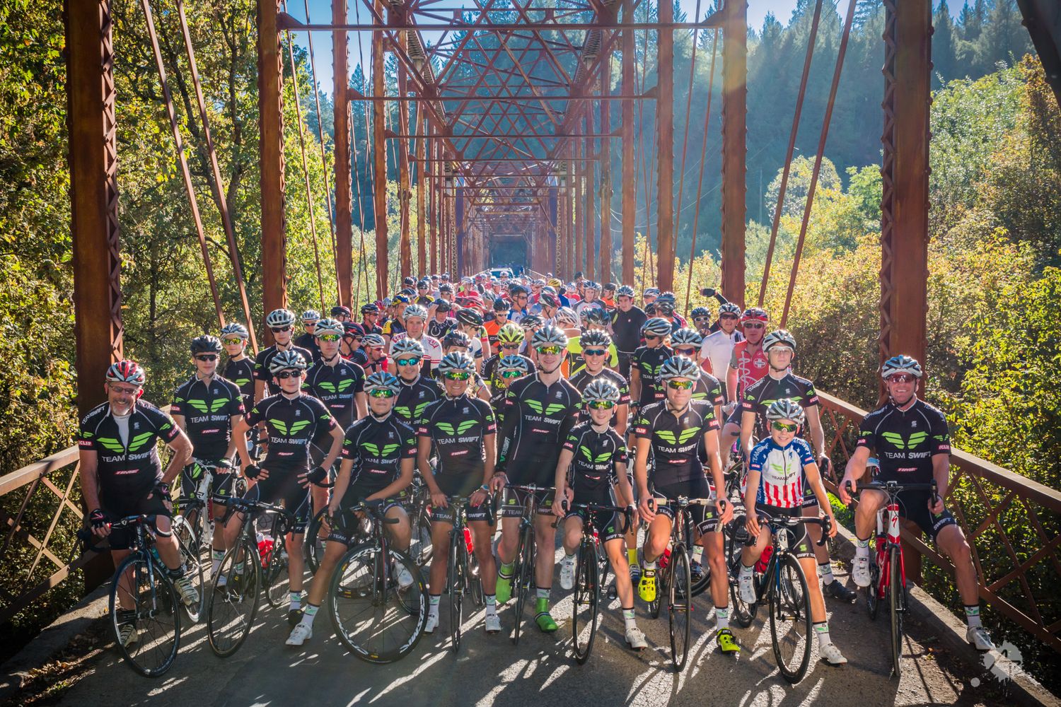 Group of cyclists gathered on a rustic bridge surrounded by autumn foliage during a racing event.