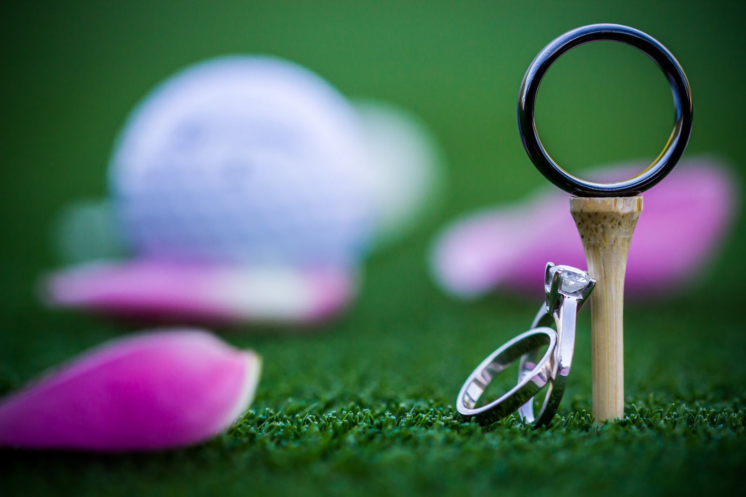 Wedding rings artistically posed on a golf tee against green background with pink flower petals.
