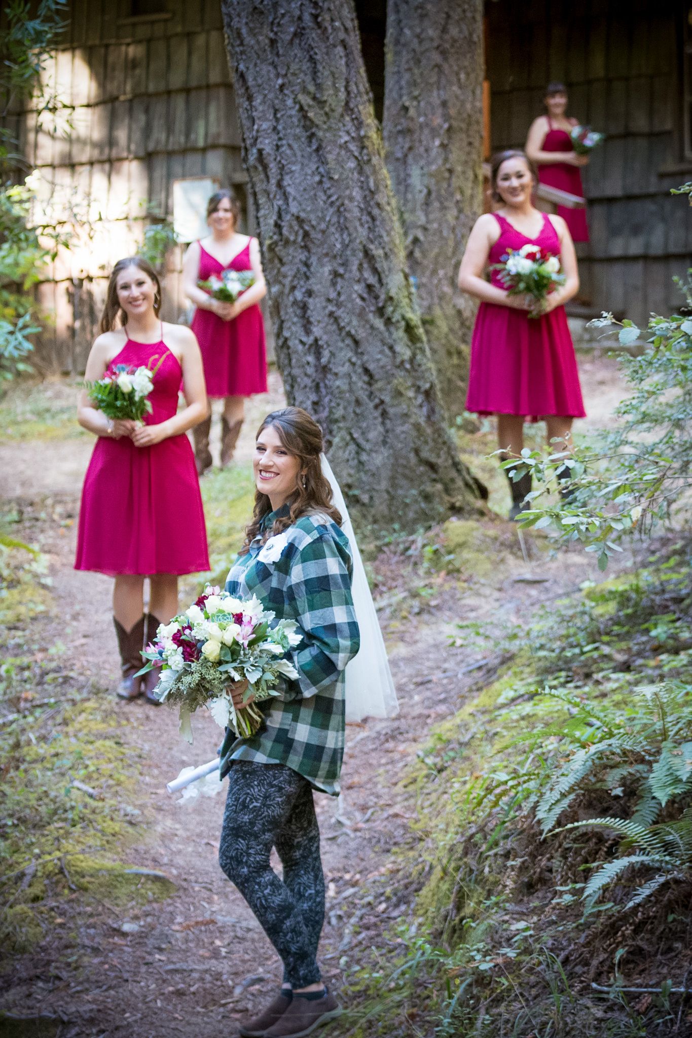 Bride in plaid with bridesmaids in red dresses holding bouquets on a forest path.