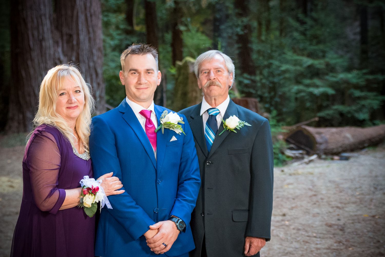 Three wedding guests in formal attire posing together against a forest backdrop.