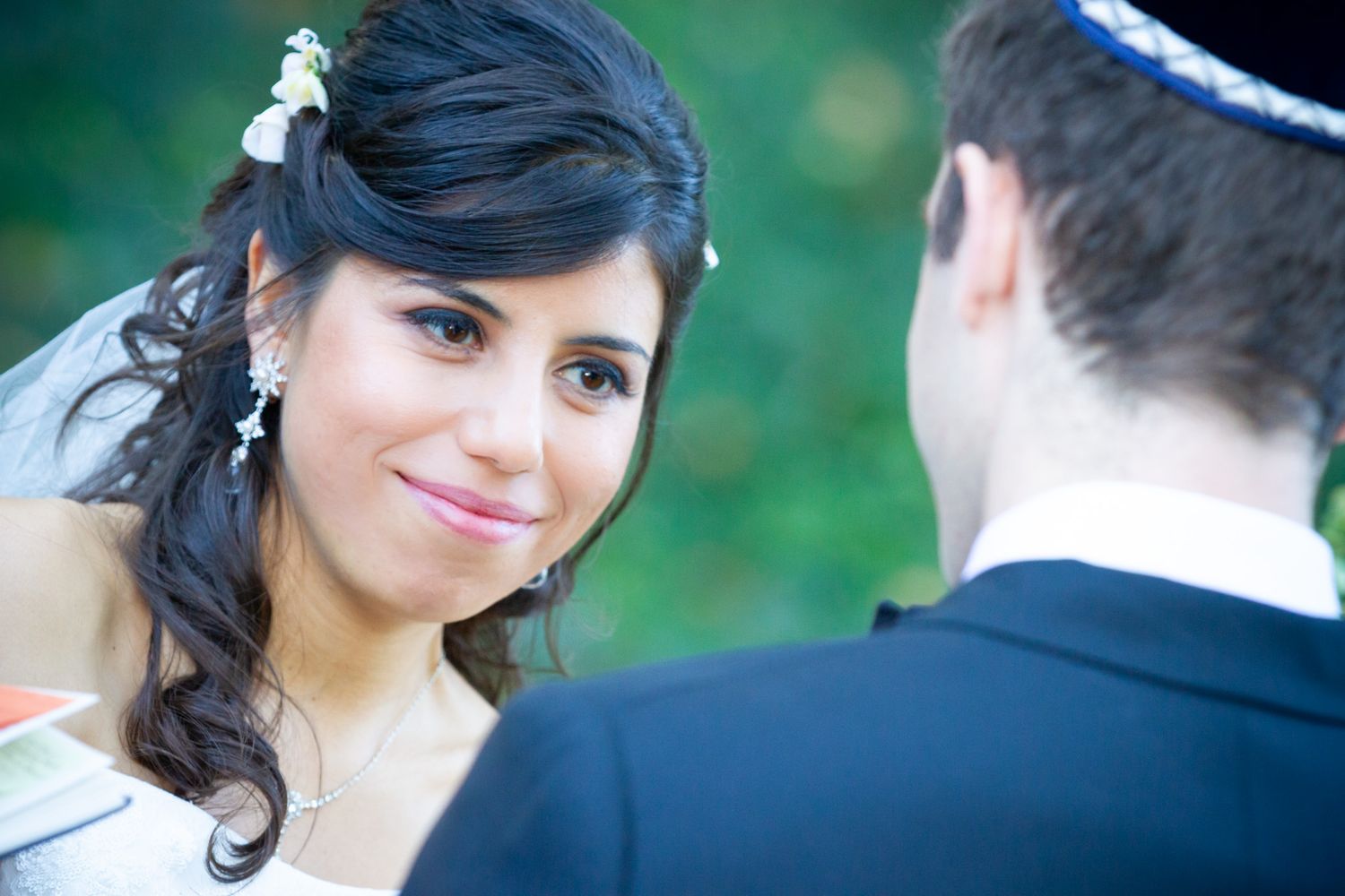A bride in a white dress and dark hair looks lovingly during an outdoor wedding ceremony.