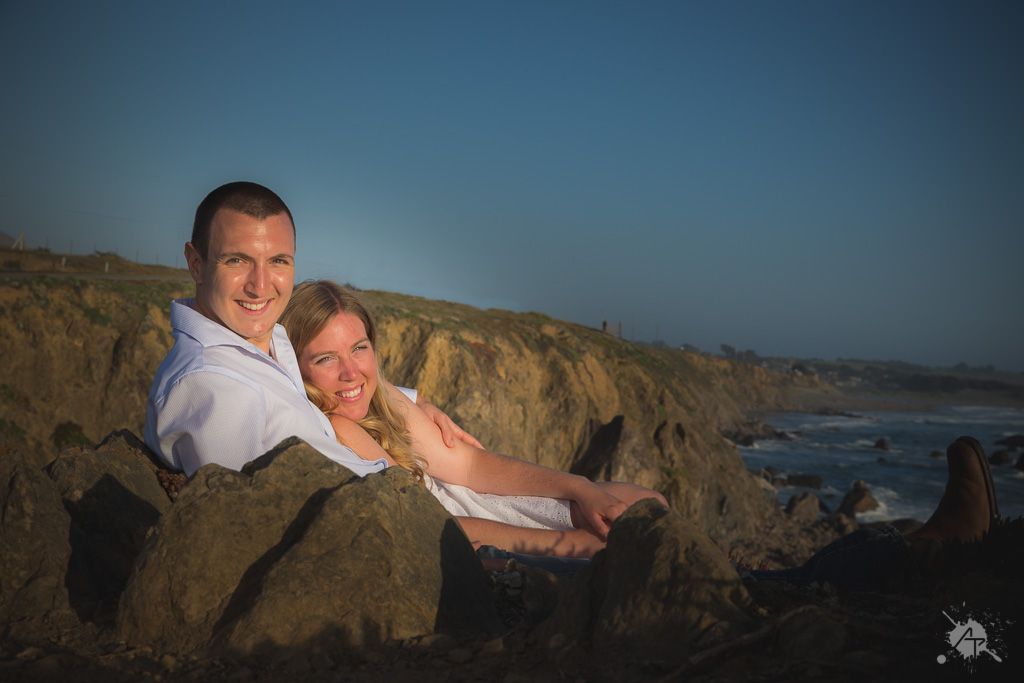 A loving couple relaxes on coastal rocks at sunset with dramatic cliffs and ocean waves in the background.