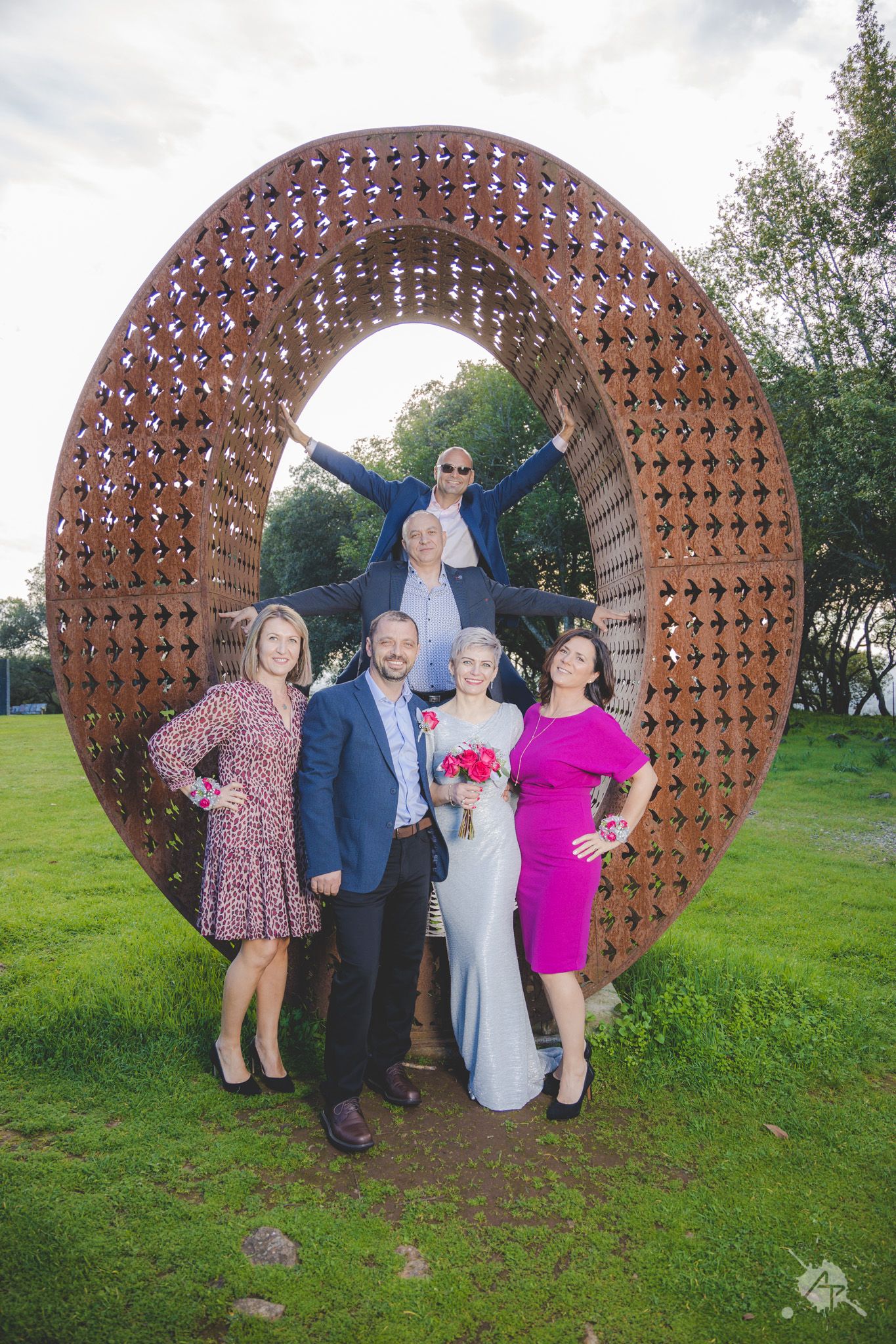 A group poses for a photo in front of a large circular metal sculpture in an outdoor garden setting.
