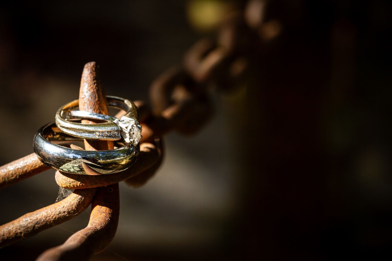Wedding rings and engagement ring artistically displayed on an antique rusted chain link in dramatic lighting.