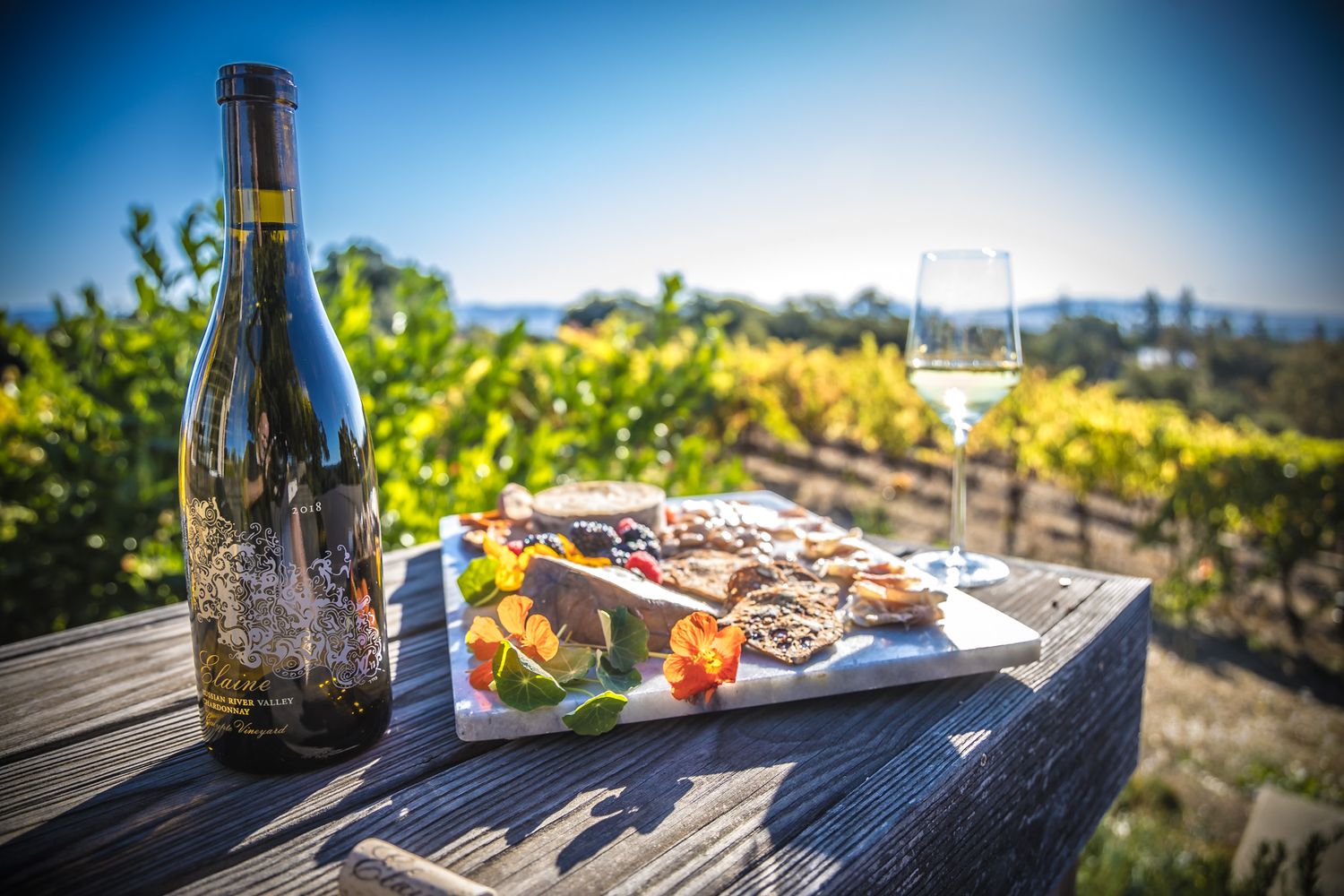 Wine bottle and food platter on rustic wooden table overlooking vineyard views.
