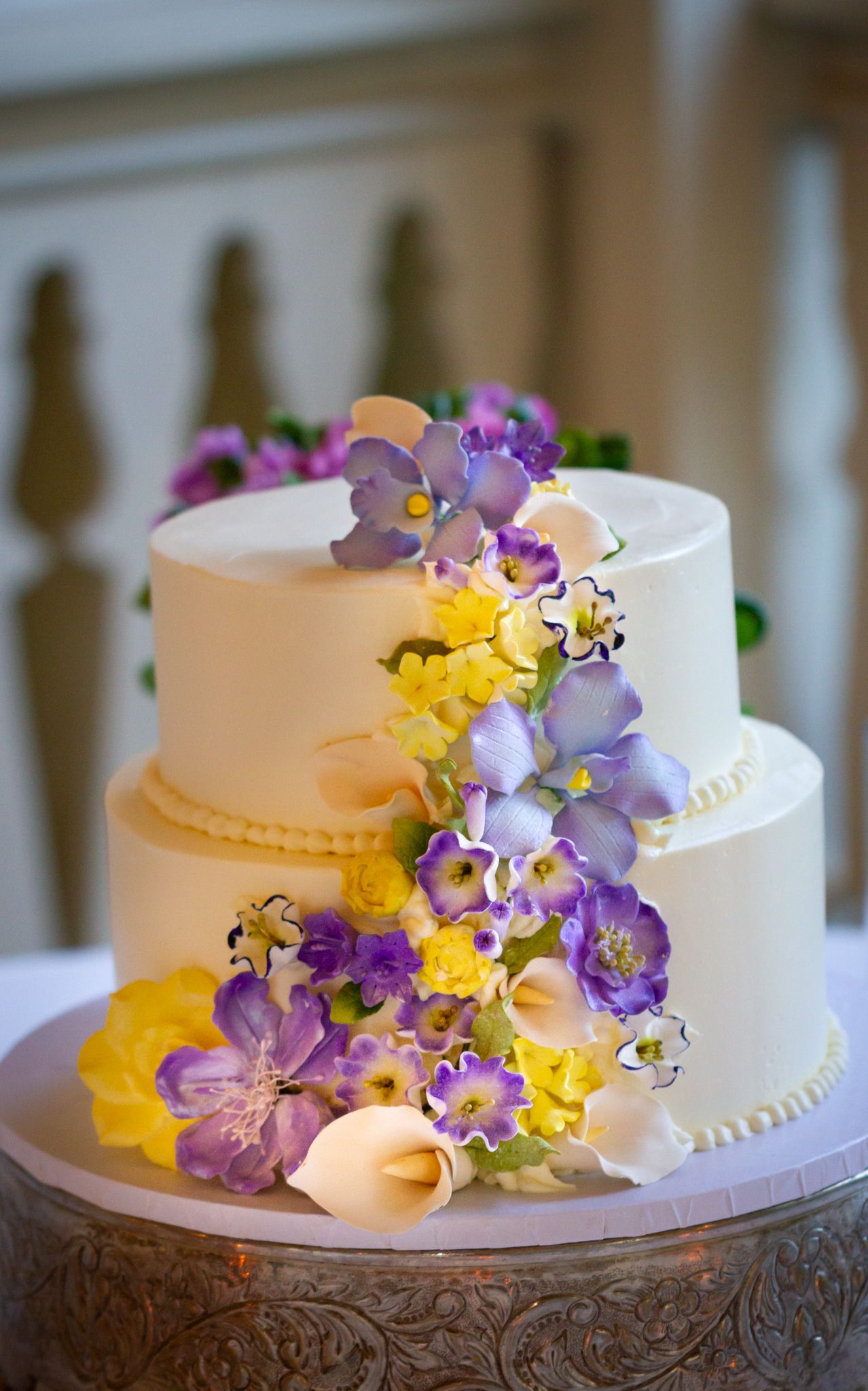 Elegant two-tier white wedding cake decorated with purple and yellow sugar flowers.