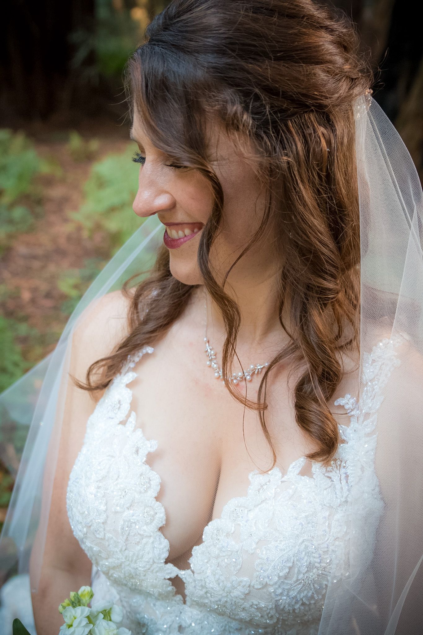 Profile shot of bride wearing lace wedding dress and veil in forest setting.