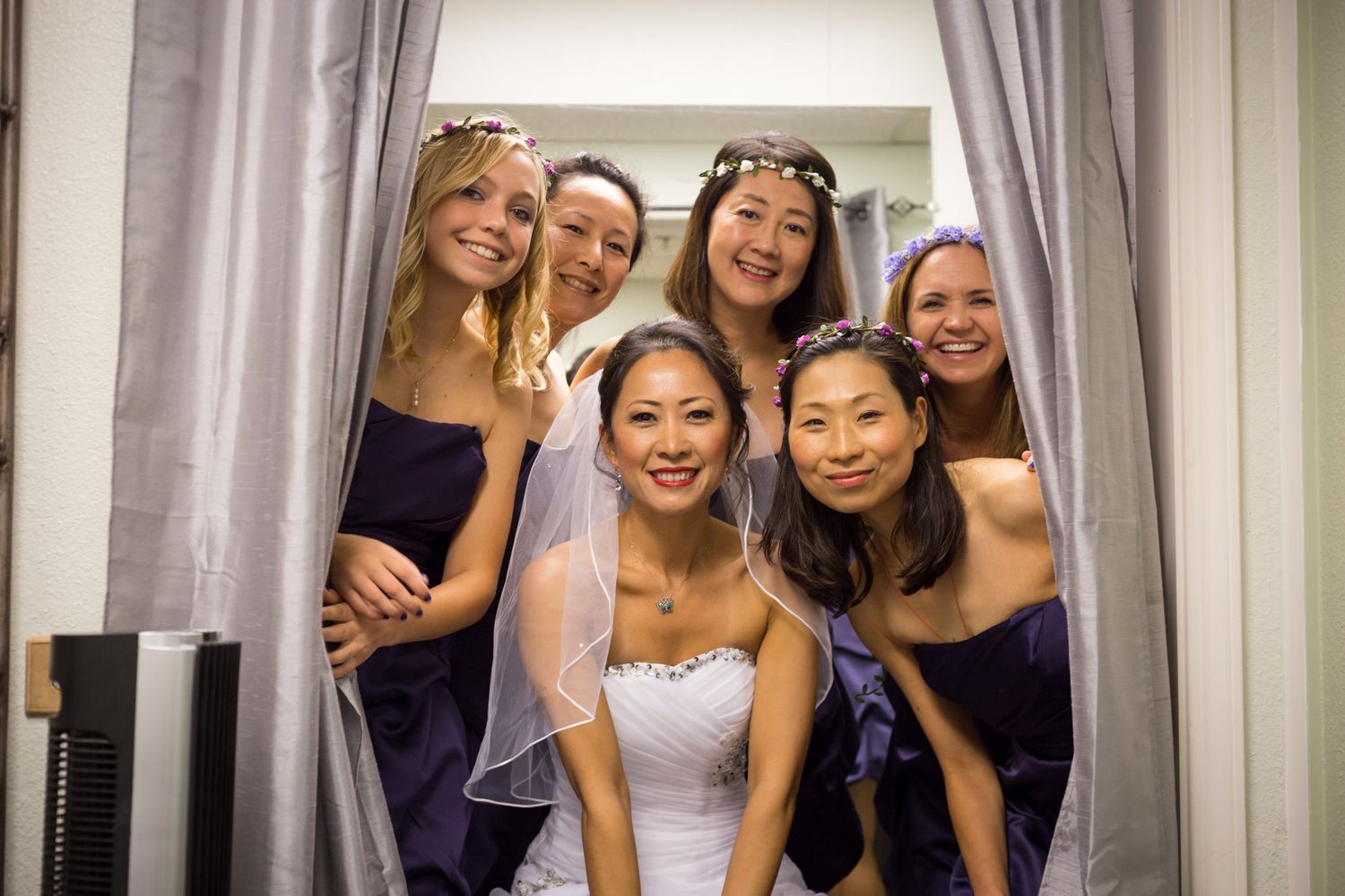 A bridal party in purple bridesmaid dresses smiles together through parted curtains at a wedding venue.