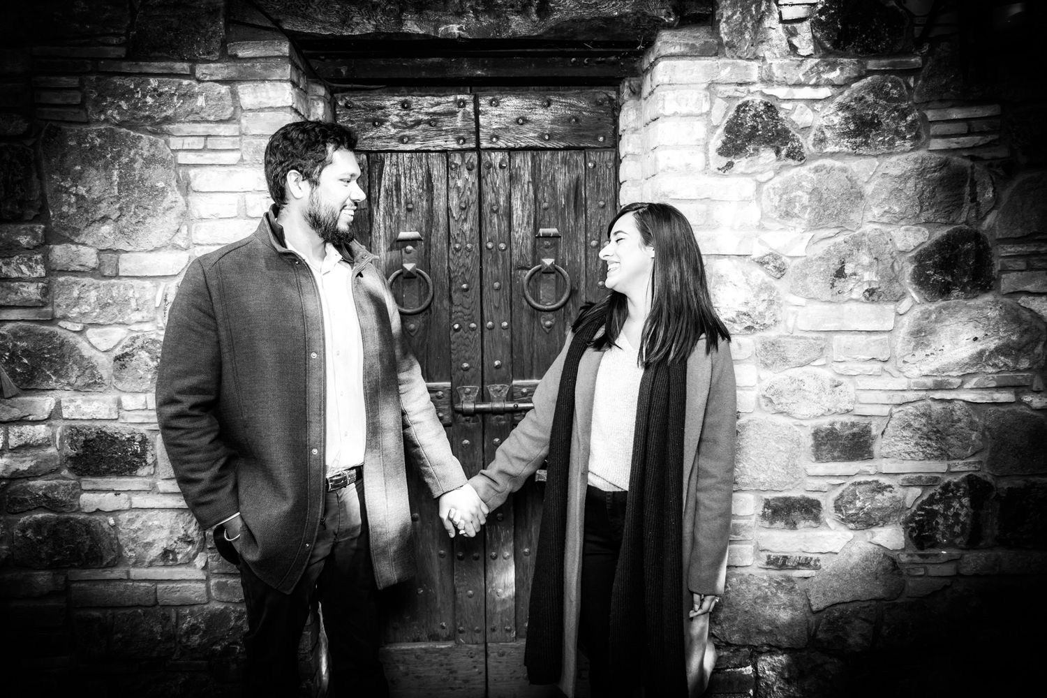 A black and white portrait of a couple holding hands in front of an old stone doorway.