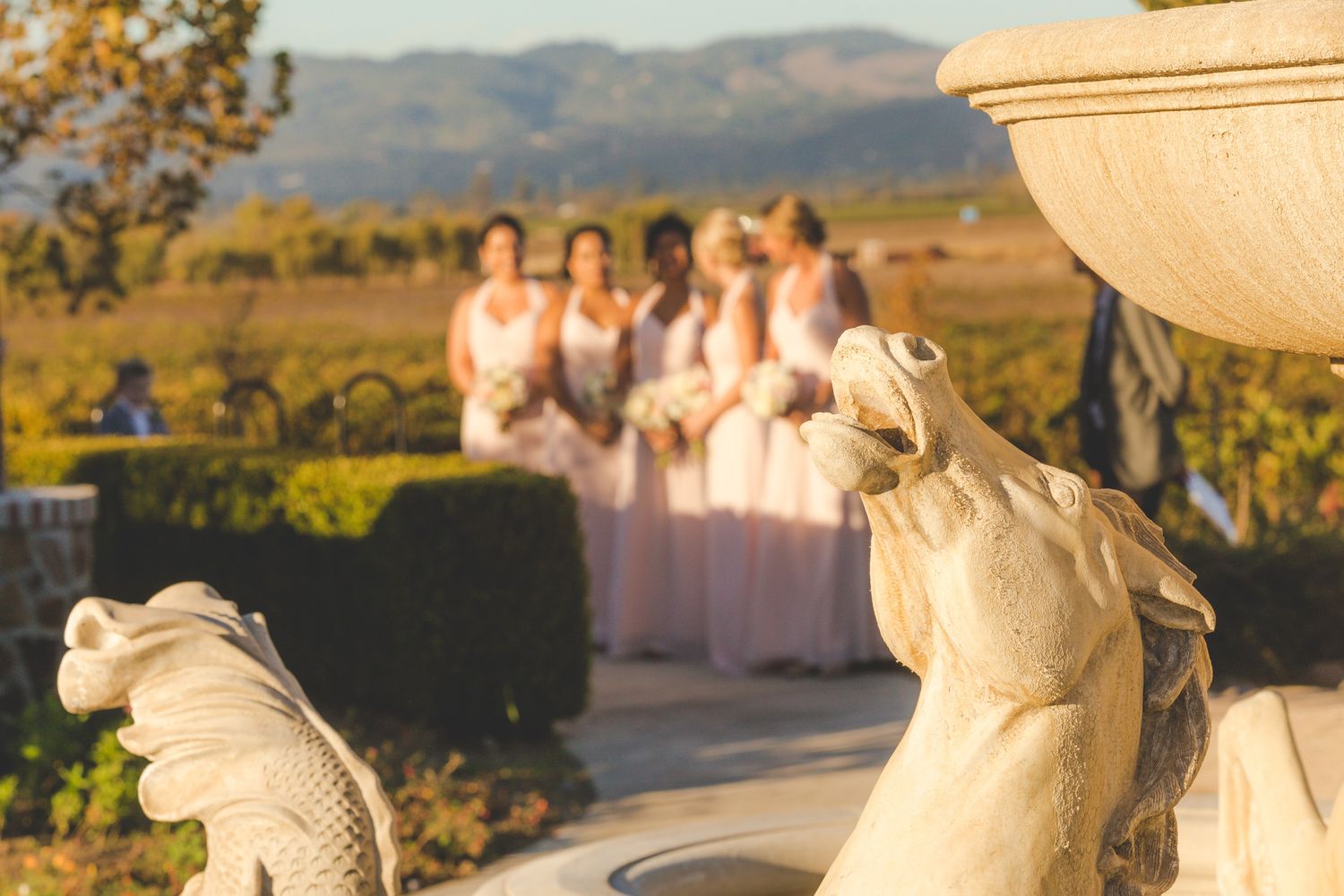 Decorative fountain statues with blurred bridesmaids in pink dresses and mountain landscape in background.
