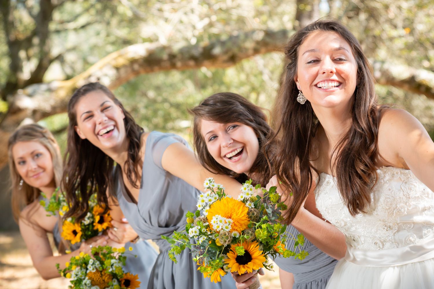 A group of people in wedding attire laughing together while holding bright yellow sunflower bouquets outdoors.