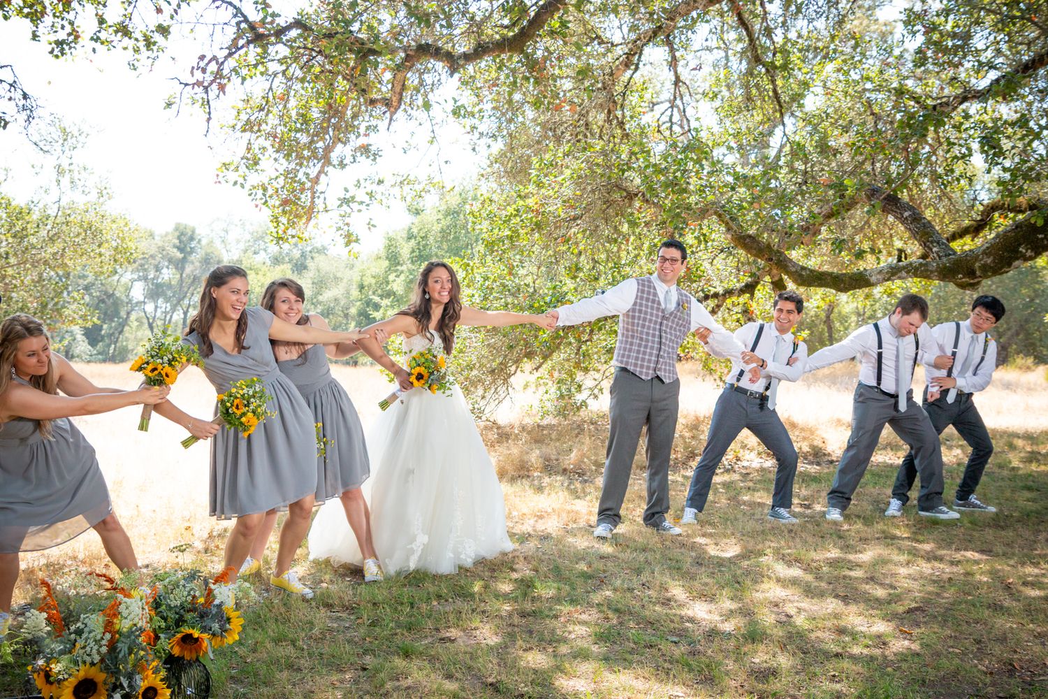 Wedding party playfully poses for a tug-of-war style photo in a sunlit outdoor setting under oak trees.