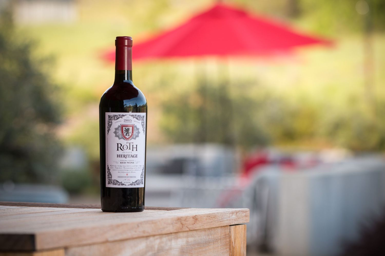 Dark wine bottle of Roth label sitting on wooden surface with red umbrella in soft focus background.