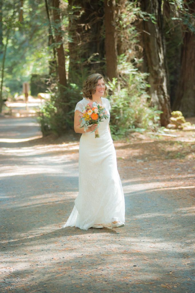 Bride carrying orange bouquet walking alone on sunlit forest path in flowing white gown.