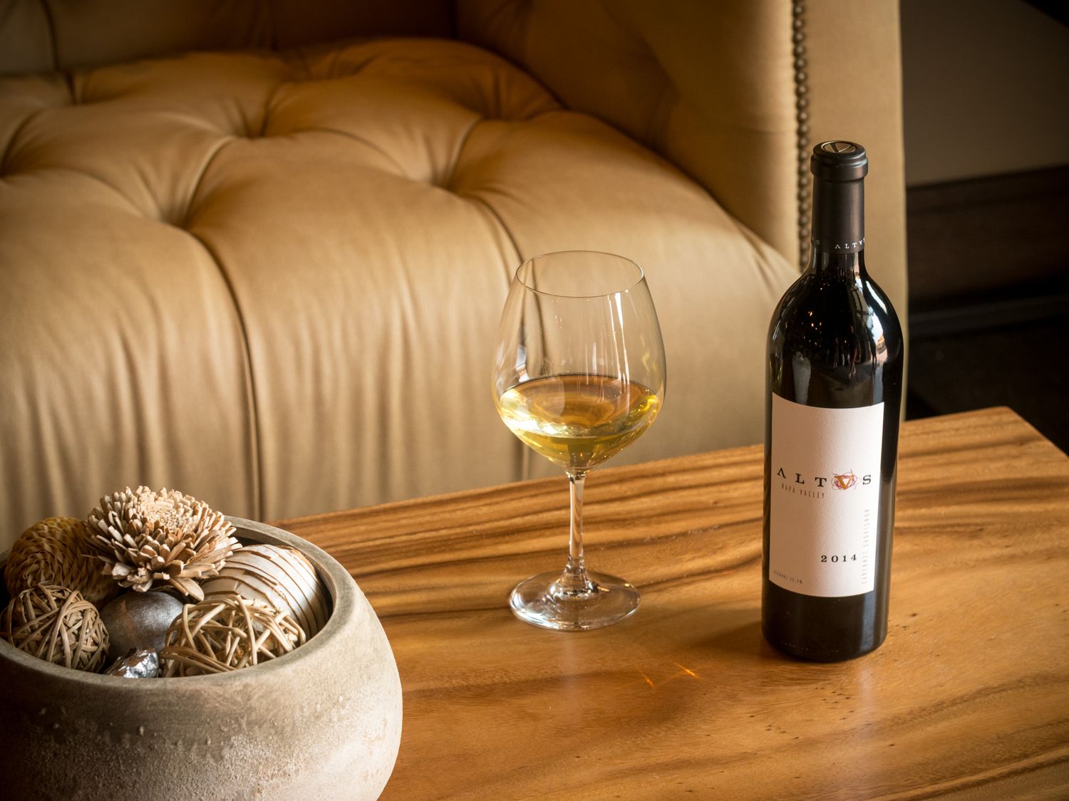 Wine bottle and glass on wooden table with tufted leather chair and decorative bowl in background.