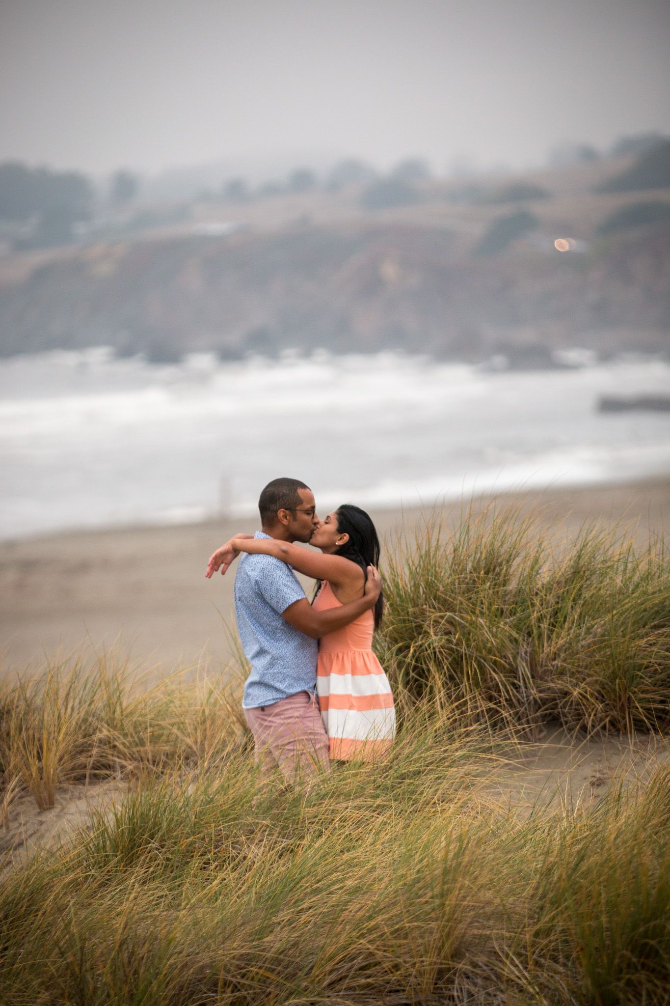 A couple embracing among beach grass with ocean views.