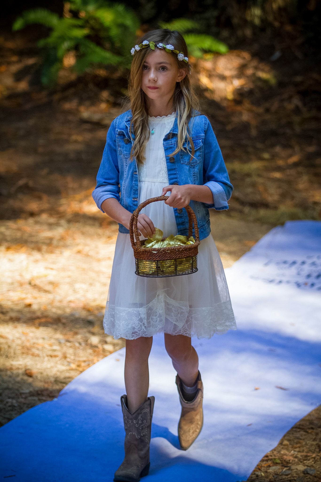 Young flower girl in white dress, denim jacket and cowboy boots holds basket while walking down blue aisle runner.