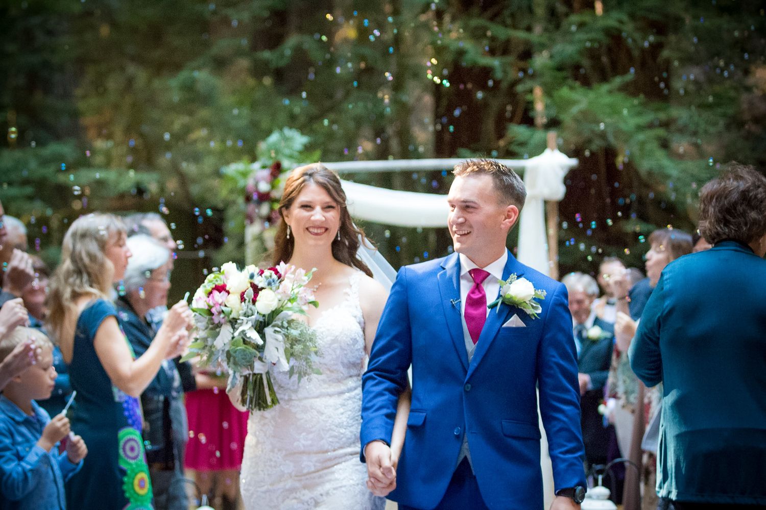 Happy newlyweds walk down the aisle while guests throw confetti in forest setting.