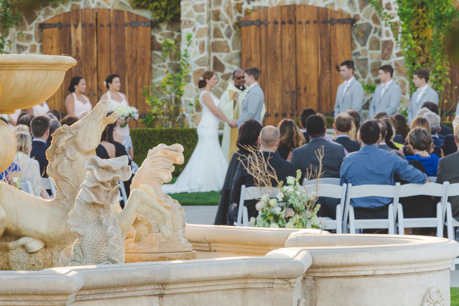 Decorative fountain with white statues serves as backdrop for an elegant outdoor wedding ceremony setup.