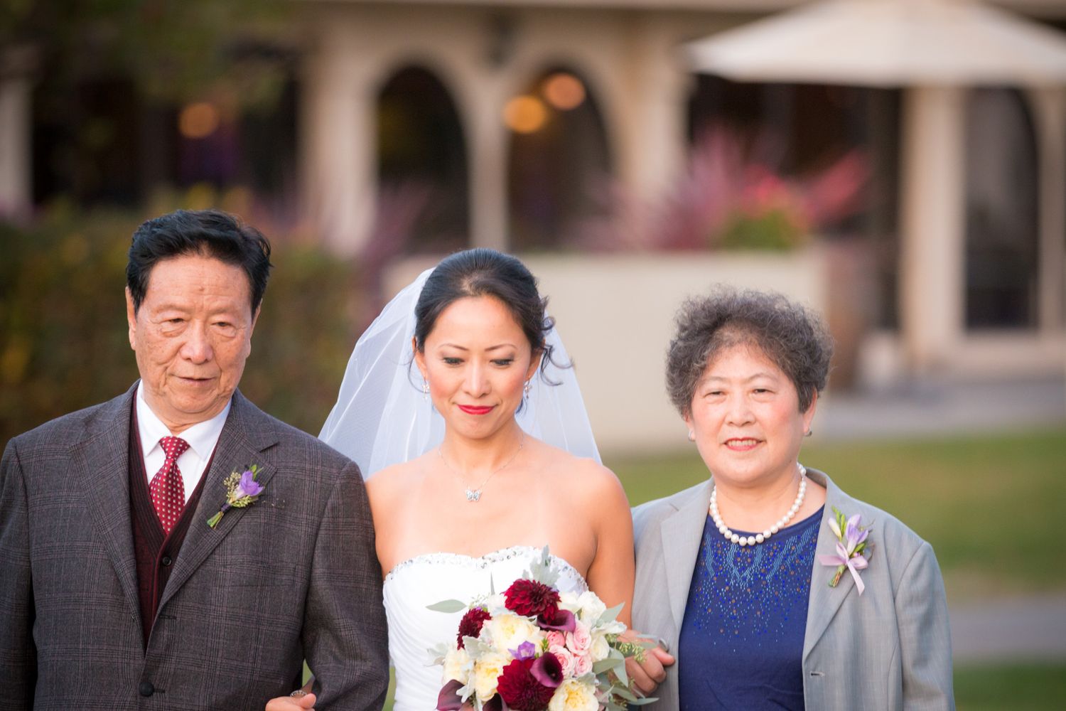 A bride in a white wedding dress holds a burgundy bouquet while standing with family members outdoors at sunset.