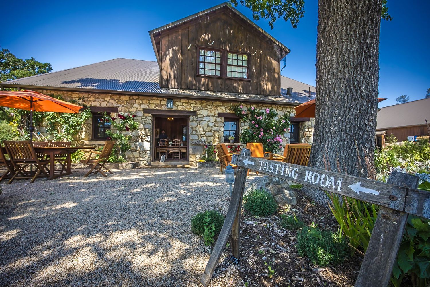 Rustic stone tasting room exterior with wooden signage and colorful garden landscaping.