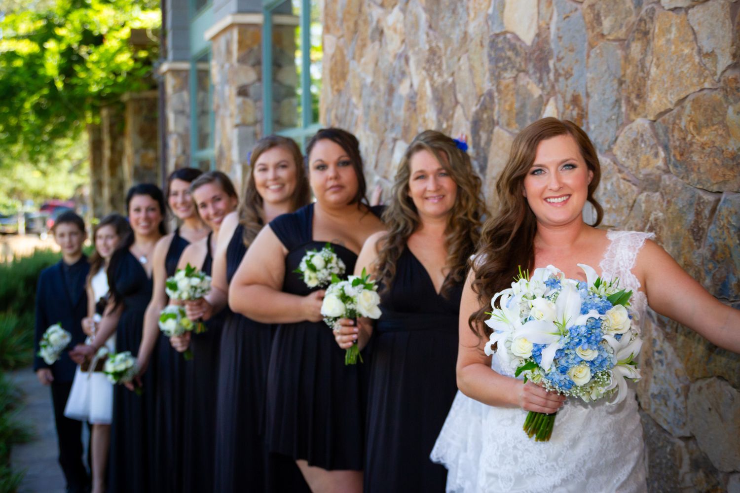 Bridesmaids in navy dresses line up along stone wall with bride holding blue bouquet.