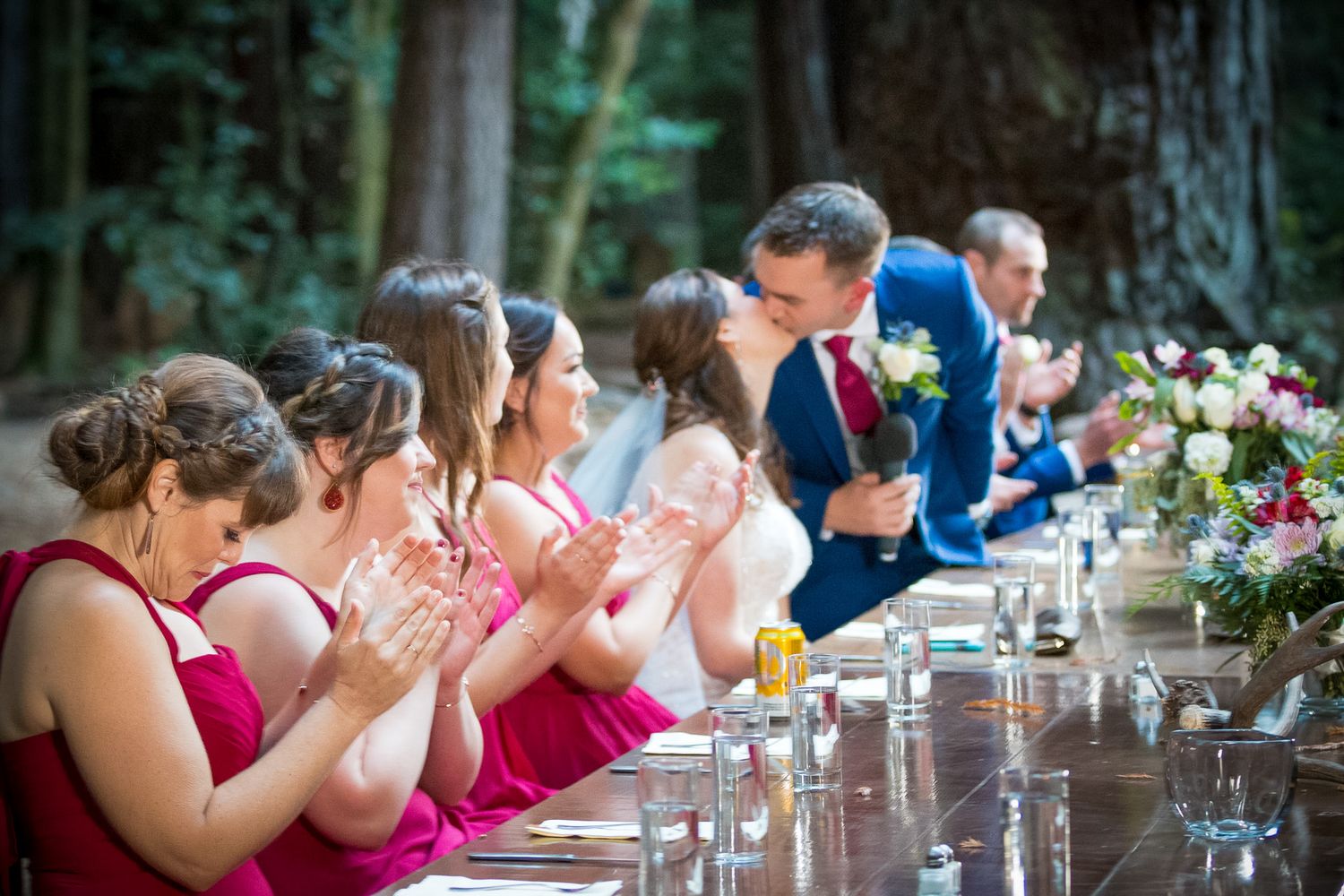 Wedding party seated at a long outdoor table in forest with bridesmaids in pink dresses applauding.