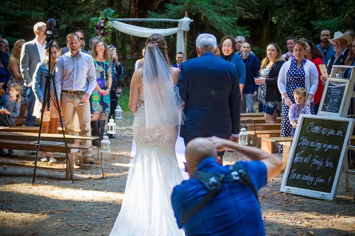 Outdoor wedding ceremony with guests seated on wooden benches in forest setting.